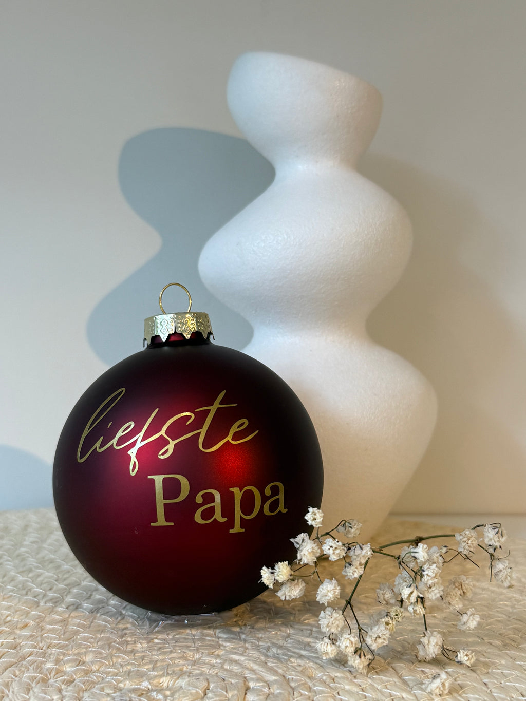 Bordeauxrode glazen kerstbal met goudkleurige tekst Liefste papa, naast een witte vaas en decoratieve droogbloemen.