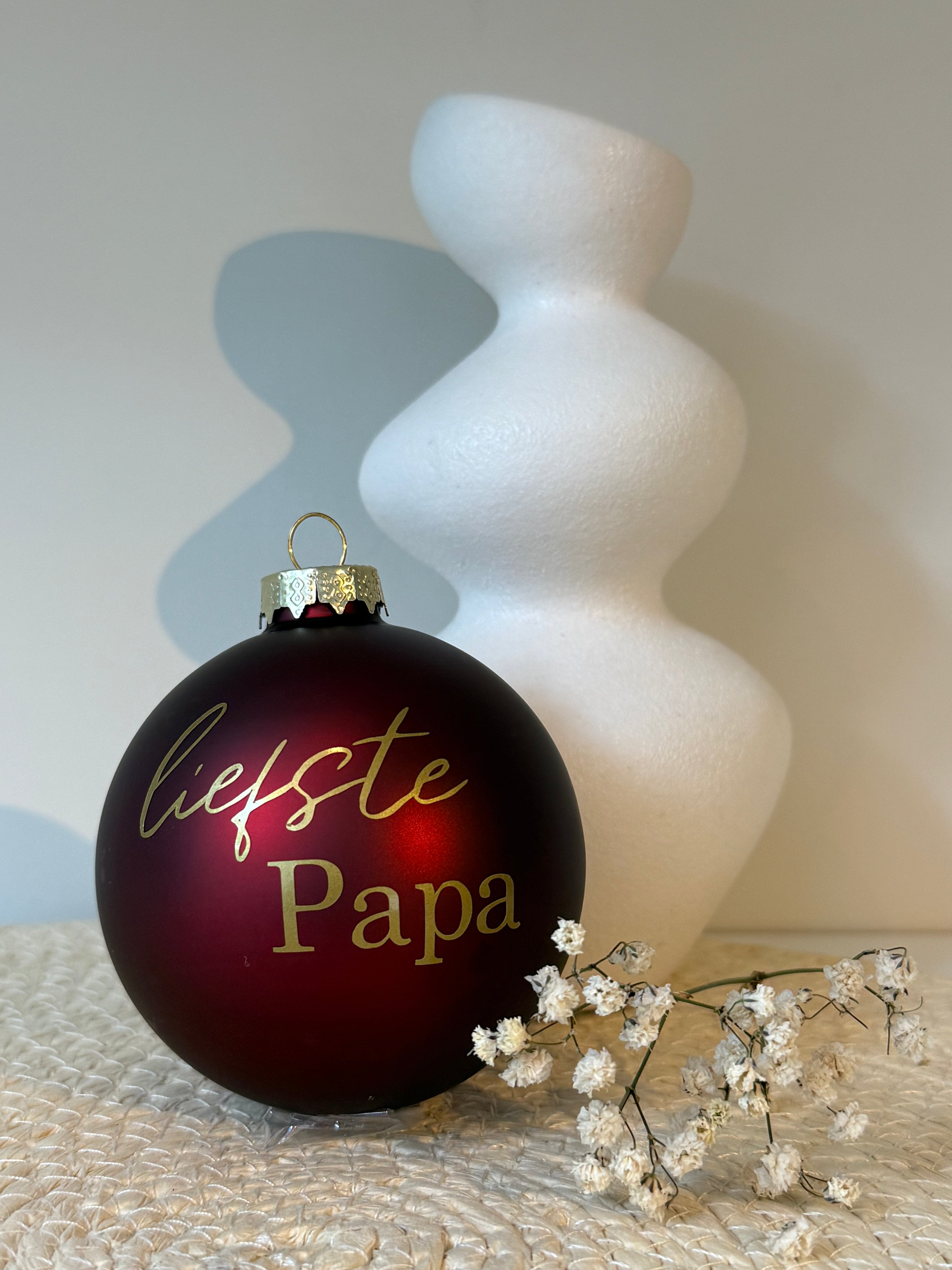 Bordeauxrode glazen kerstbal met goudkleurige tekst Liefste papa, naast een witte vaas en decoratieve droogbloemen.