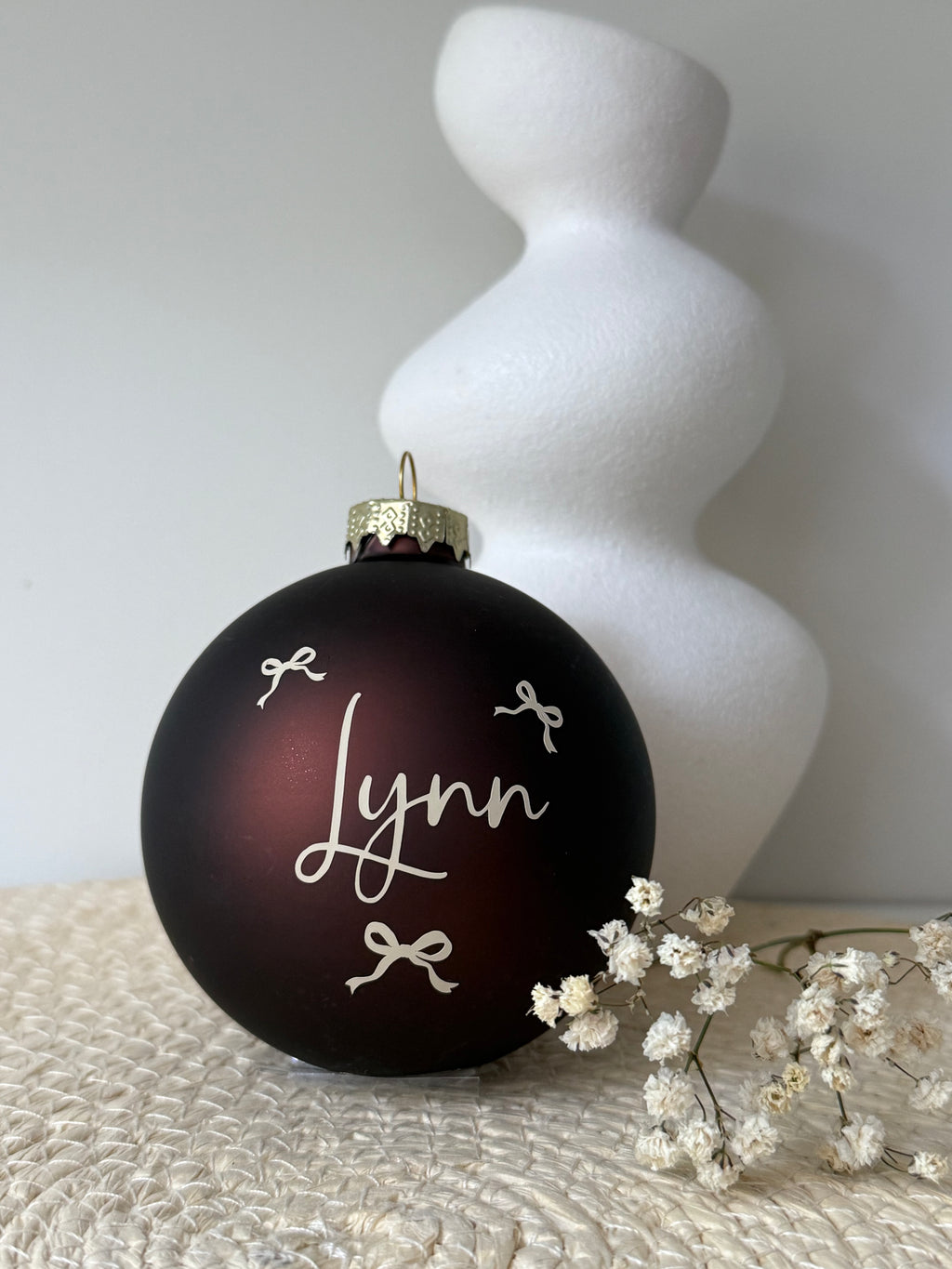 Bruine kerstbal met een warme bordeauxrode ondertoon, gepersonaliseerde naam en sierlijke strikjes, naast een witte vaas met decoratie.