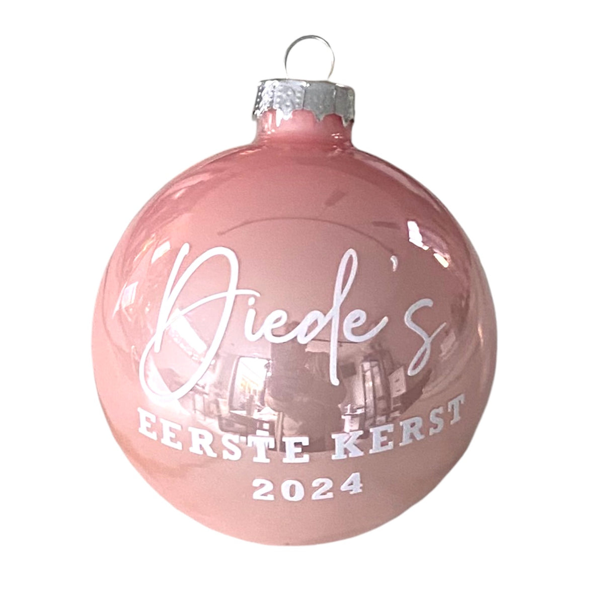 Roze glazen kerstbal met gepersonaliseerde witte naamopdruk, tekst Eerste kerst en jaartal, tegen een witte achtergrond.