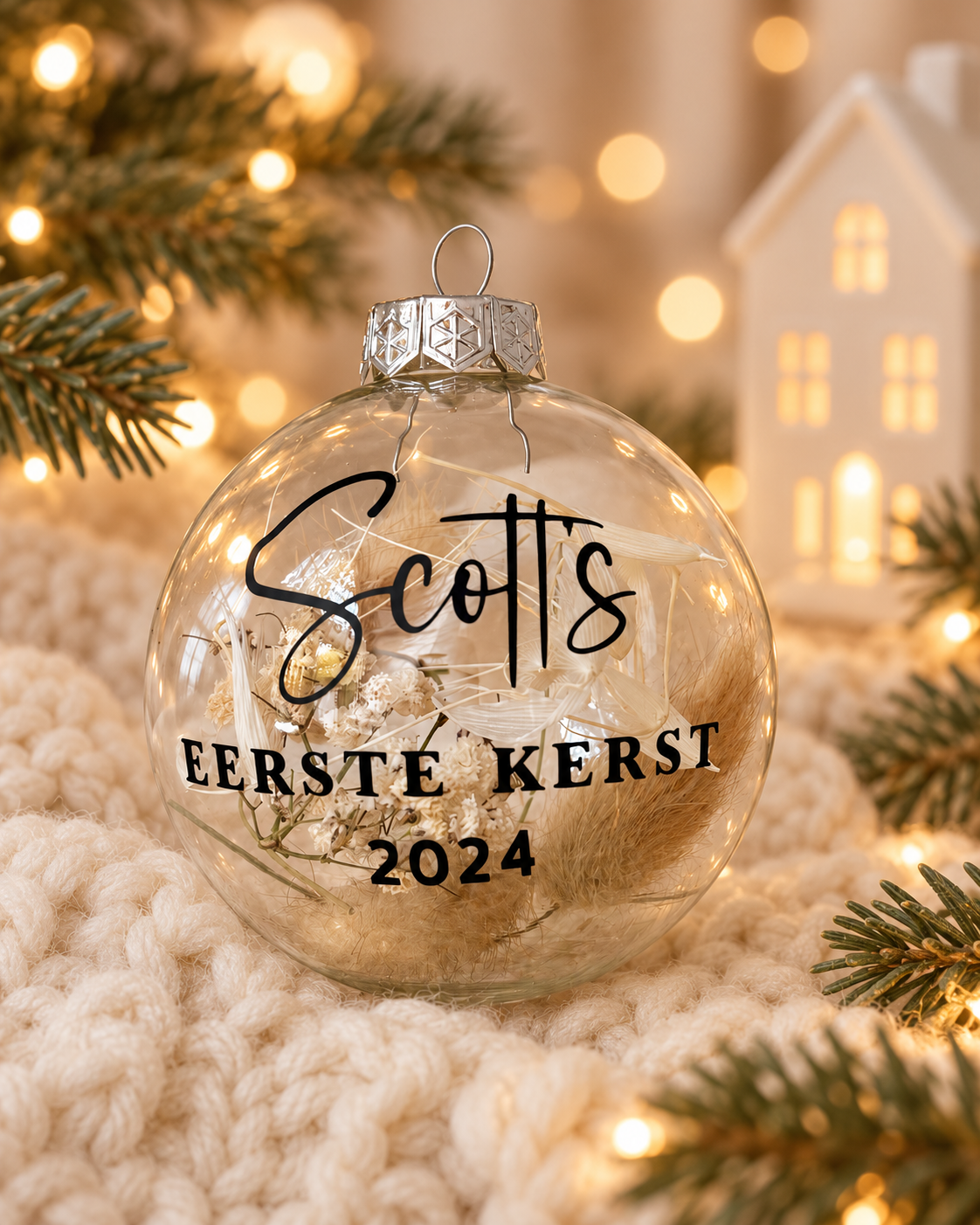 Transparante glazen kerstbal met droogbloemen, zwarte naamopdruk en tekst Eerste kerst, in een warme kerstsetting.