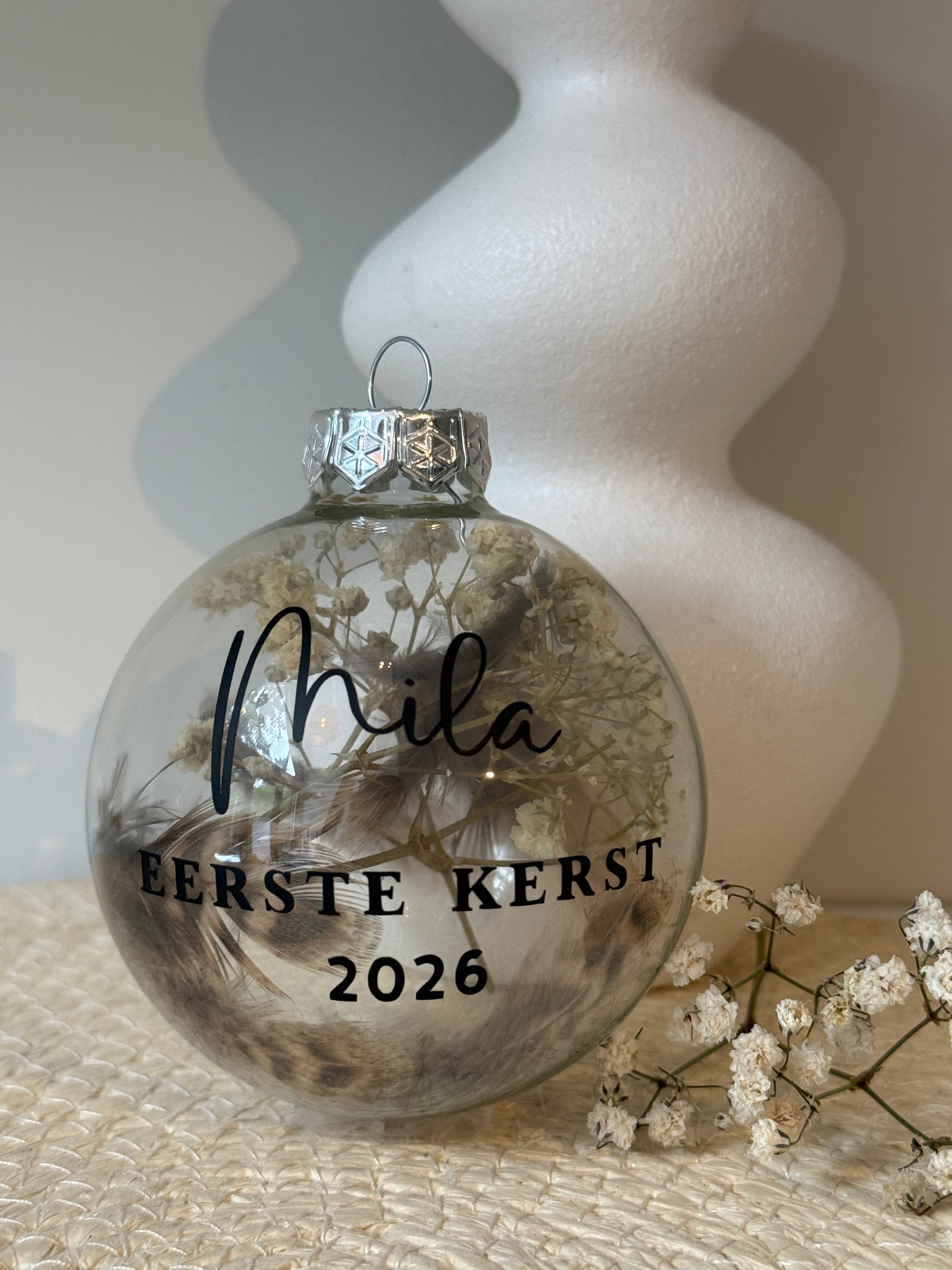 Detailfoto van een transparante glazen kerstbal met droogbloemen, gepersonaliseerde zwarte naamopdruk en jaartal.