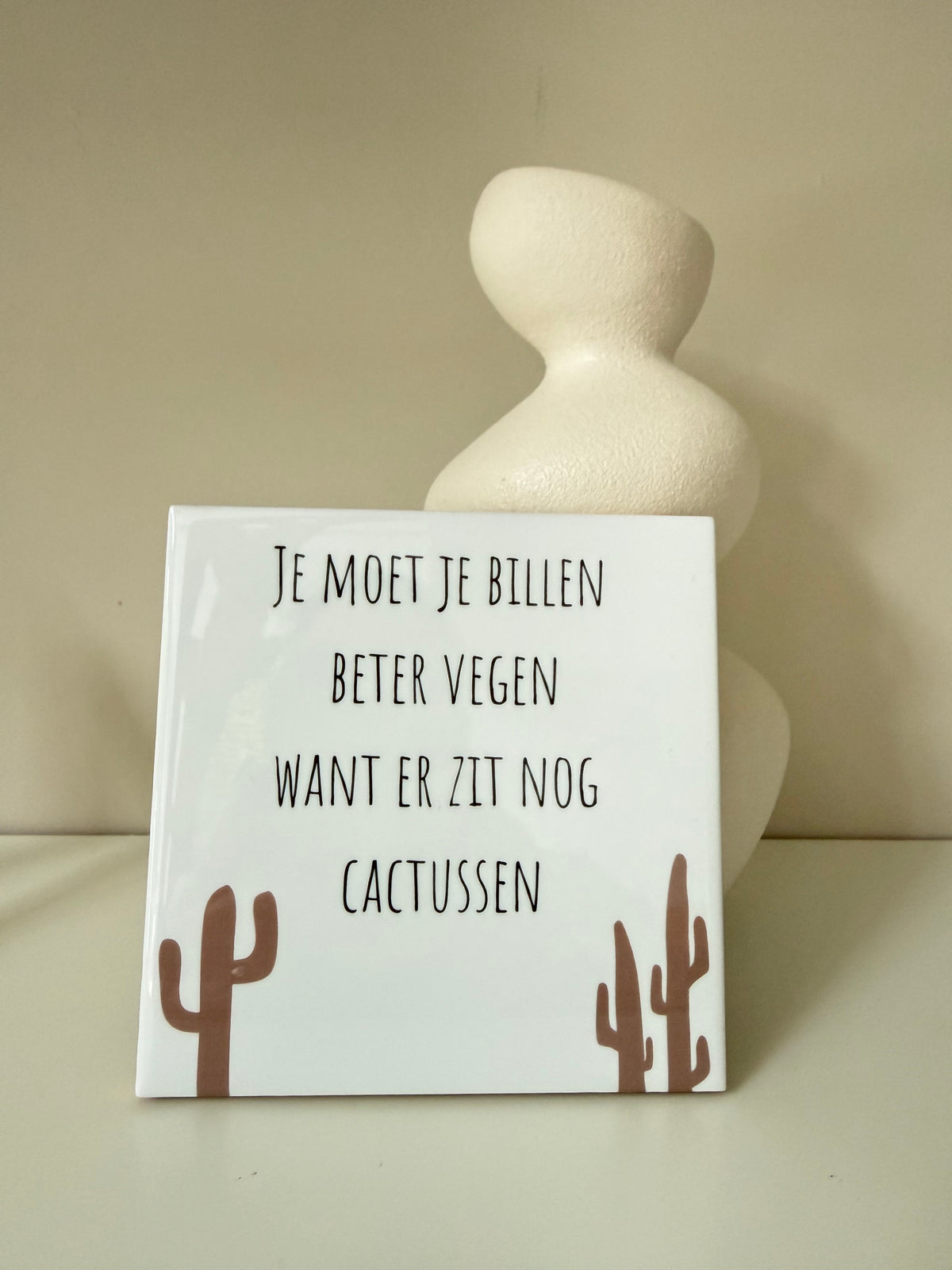 Wit tegeltje met grappige tekst over billen vegen en cactusillustraties, op een houten houdertje naast een witte vaas.
