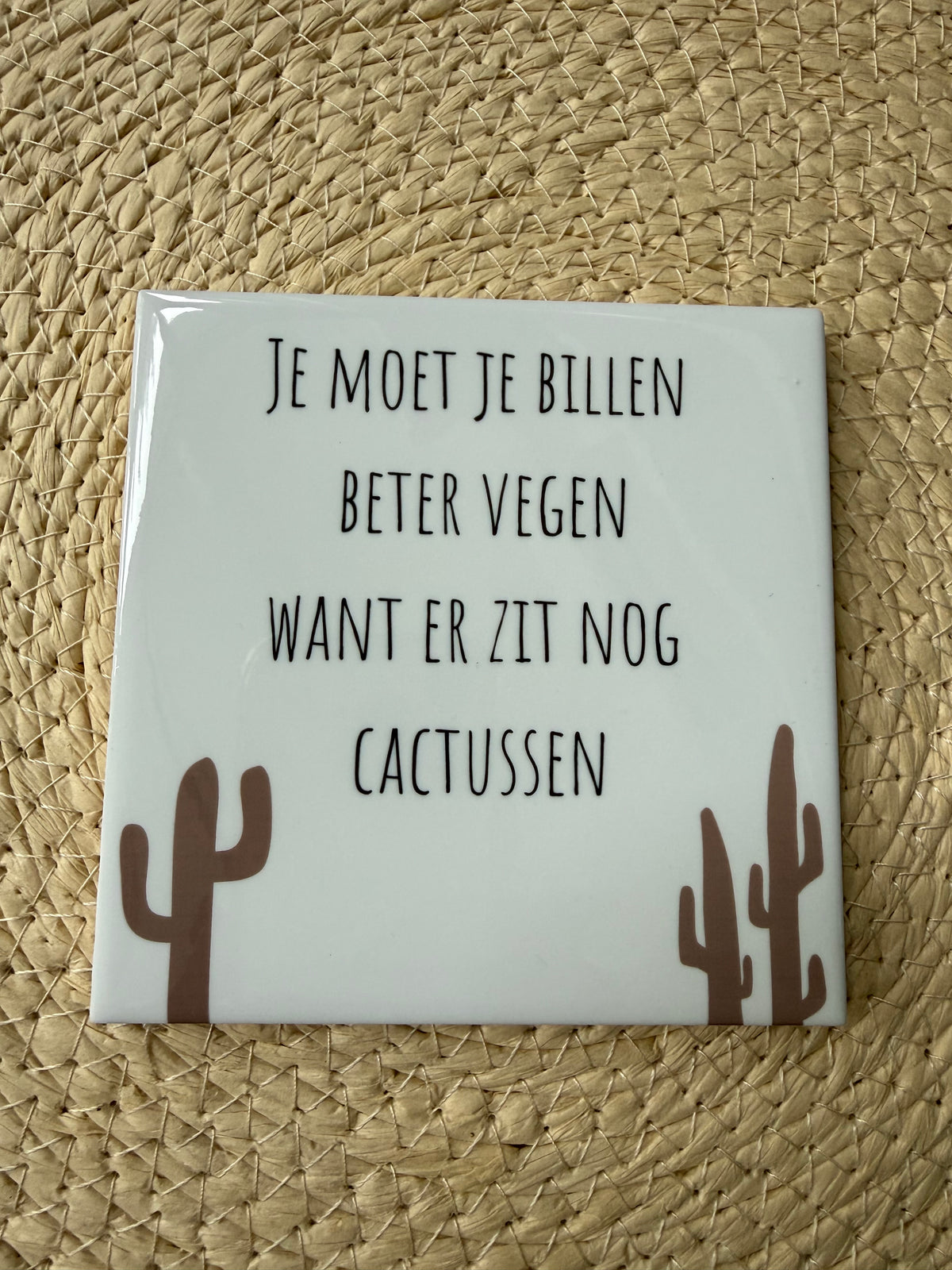 Wit keramieken WC-tegeltje met humoristische tekst en bruine cactusillustraties, liggend op een  rieten ondergrond.