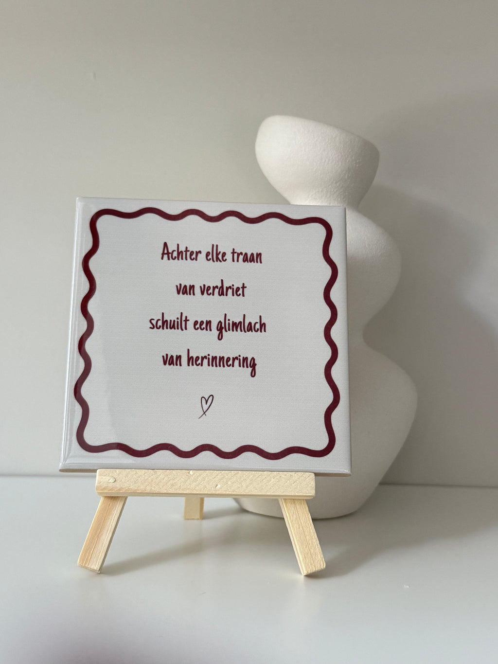 Herinneringstegeltje met de tekst 'Achter elke traan van verdriet schuilt een glimlach van herinnering' op houten standaard naast een witte vaas.