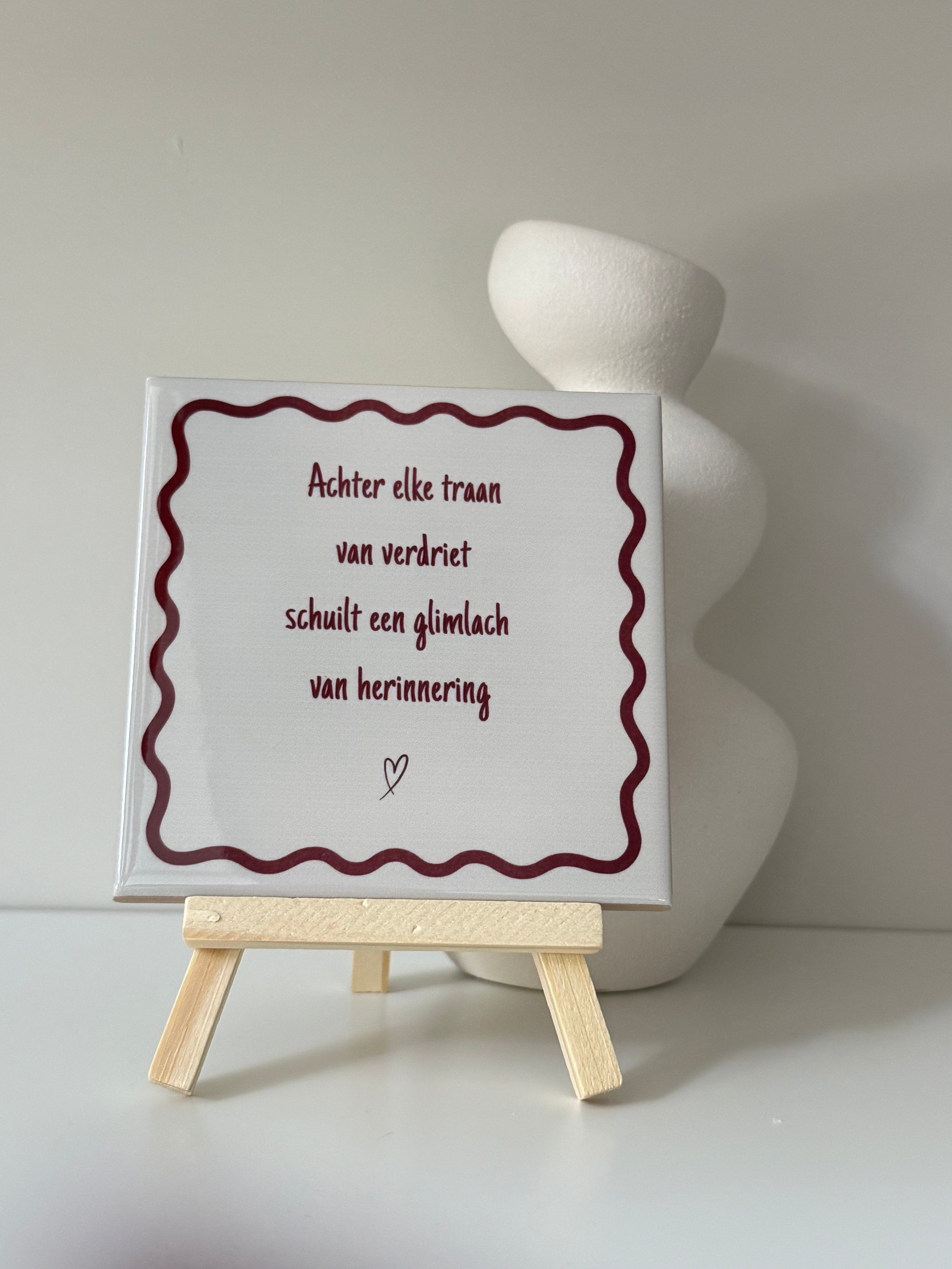 Herinneringstegeltje met de tekst 'Achter elke traan van verdriet schuilt een glimlach van herinnering' op houten standaard naast een witte vaas.
