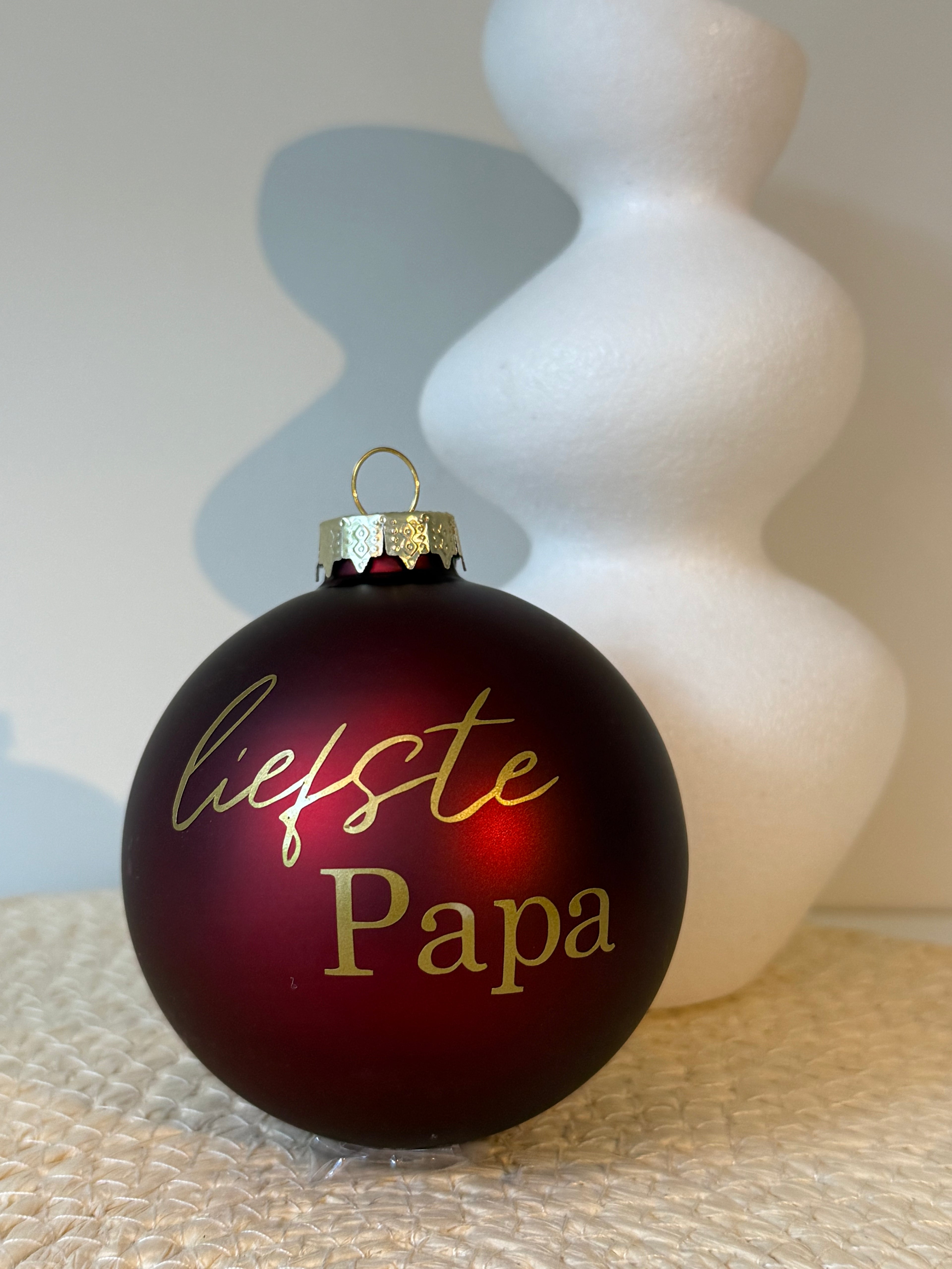 Matte bordeauxrode glazen kerstbal met gouden tekst Liefste papa, op een beige ondergrond met een witte vaas op de achtergrond.