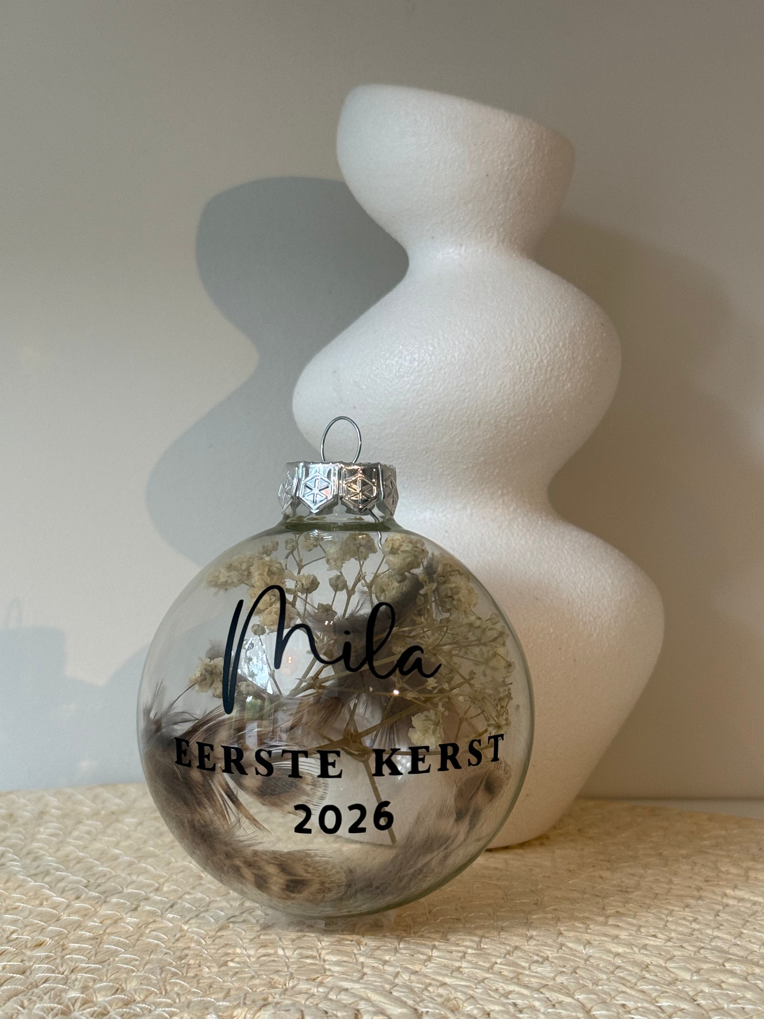 Gepersonaliseerde glazen kerstbal met droogbloemen en zwarte tekst Eerste kerst, op een originele productfoto.