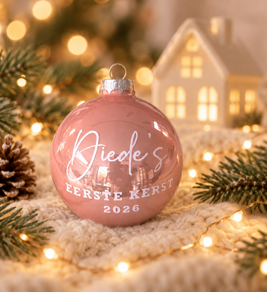 Roze glazen kerstbal met gepersonaliseerde witte naamopdruk, tekst Eerste kerst en jaartal, tegen een witte achtergrond.
