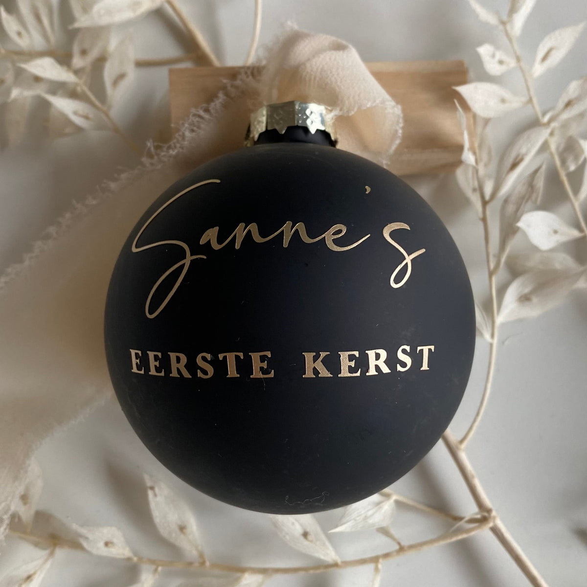 Gepersonaliseerde matzwarte glazen kerstbal met goudkleurige opdruk voor de eerste kerst, gefotografeerd in een sfeervolle kerstsetting.
Kopiëren